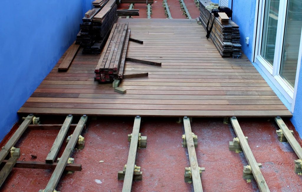 Wooden deck installation in progress with boards partially laid over a metal frame on a reddish outdoor surface.